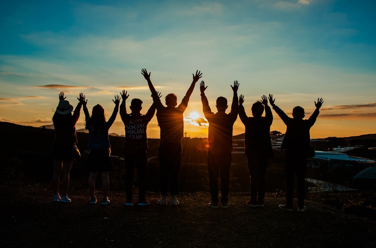 about-us-02 A diverse group of friends raises their arms in celebration against a vibrant sunset backdrop.